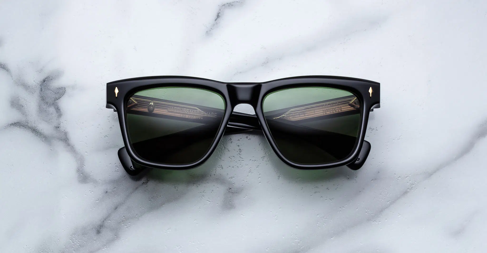 A pair of Jacques Marie Mage Lankaster black rectangular sunglasses with dark green lenses rests on a white marble surface.