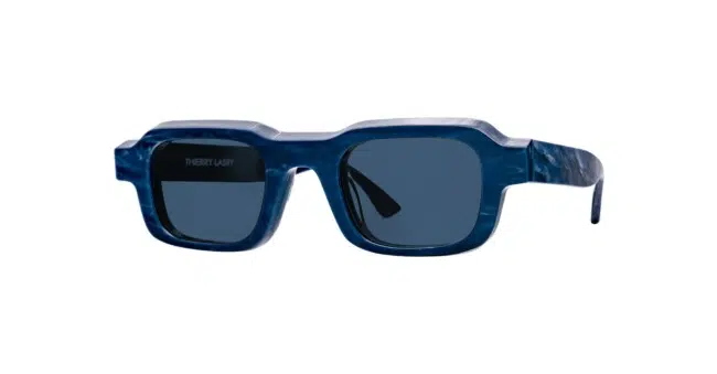 The Thierry Lasry X Reese Cooper Flexxxy sunglasses feature a rectangular shape, thick marbled blue frames, and dark tinted lenses, displayed against a white background.