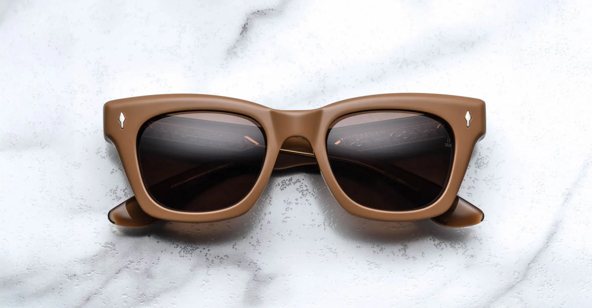 The Jacques Marie Mage Dealan X Umit Benan brown square-framed sunglasses with dark lenses are displayed on a white marble surface.
