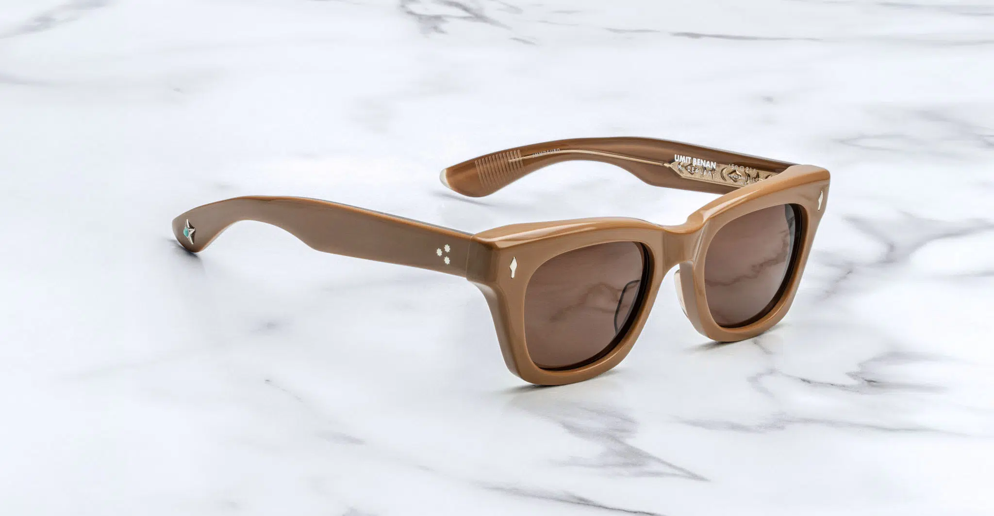 The Jacques Marie Mage Dealan X Umit Benan are brown rectangular sunglasses with thick frames and dark lenses, shown on a white marble surface. The temples have small decorative details near the hinges.