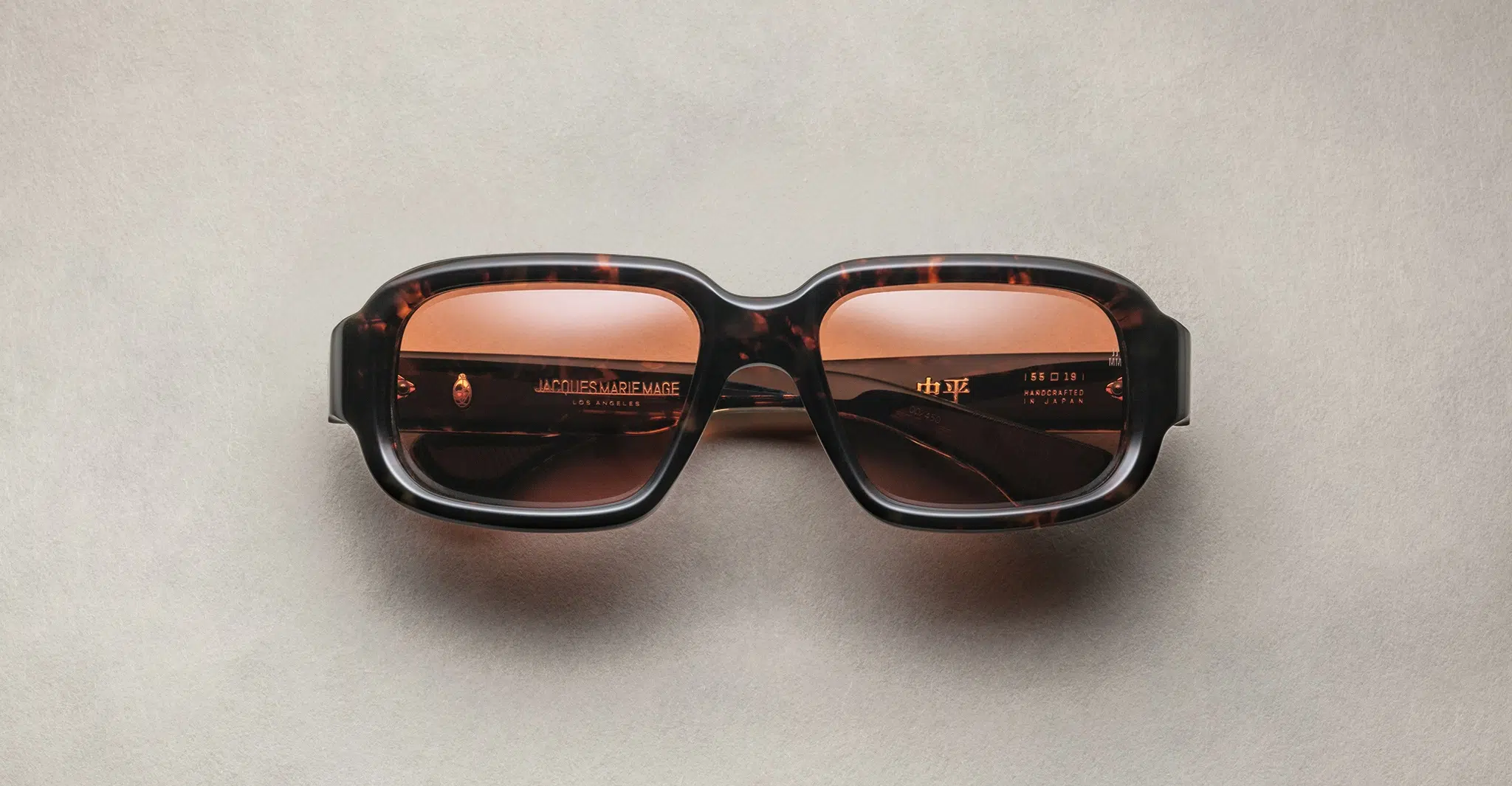 Jacques Marie Mage Nakahira rectangular tortoiseshell sunglasses with thick frames and brown tinted lenses, shown on a light beige surface.