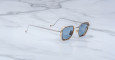 The Jacques Marie Mage Atkins sunglasses, featuring gold and clear frames with blue-tinted lenses, are displayed on a white marble surface with gray veining.