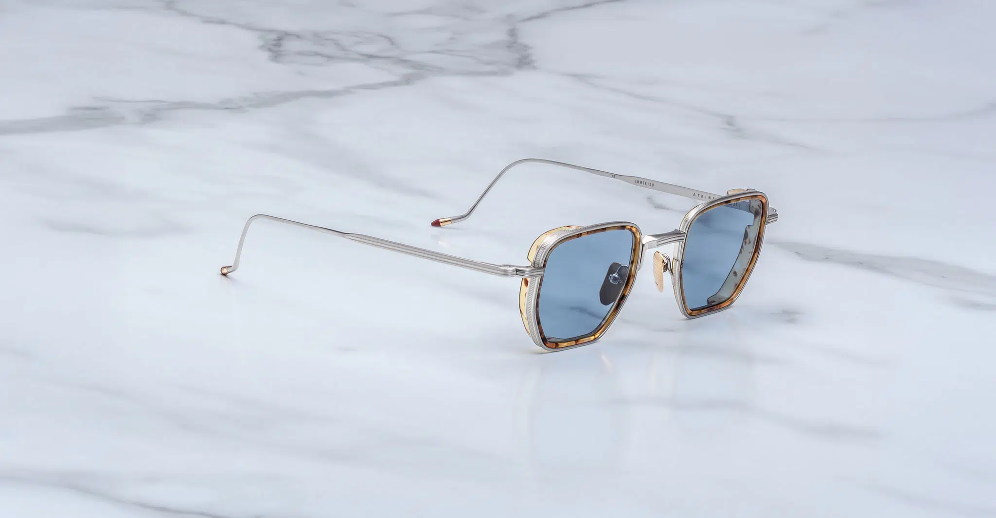 The Jacques Marie Mage Atkins sunglasses, featuring gold and clear frames with blue-tinted lenses, are displayed on a white marble surface with gray veining.