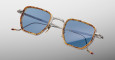 The Jacques Marie Mage Atkins eyeglasses feature square silver metal frames with tortoiseshell-patterned rims, blue tinted lenses, and red-tipped temples, set against a smooth white-to-gray gradient background.