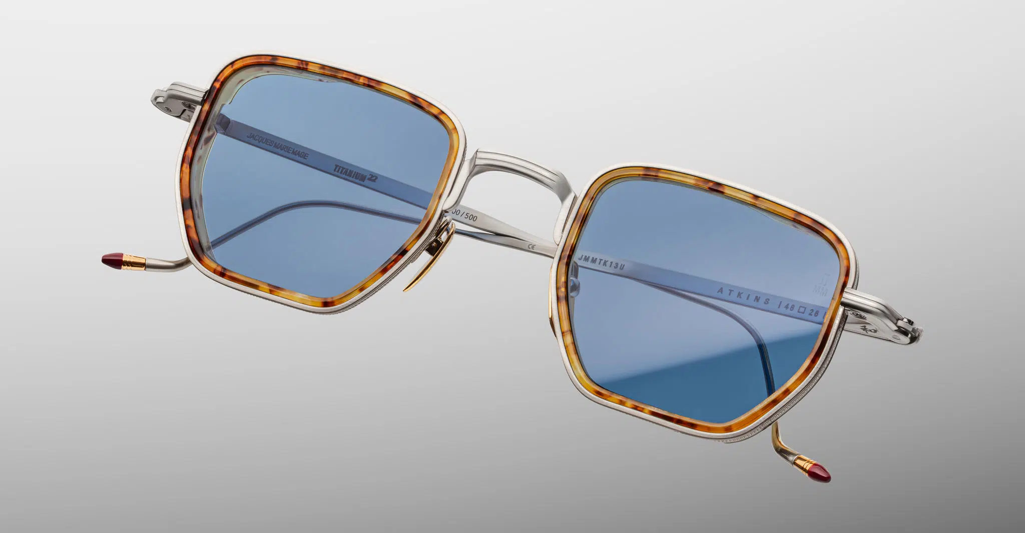 The Jacques Marie Mage Atkins eyeglasses feature square silver metal frames with tortoiseshell-patterned rims, blue tinted lenses, and red-tipped temples, set against a smooth white-to-gray gradient background.