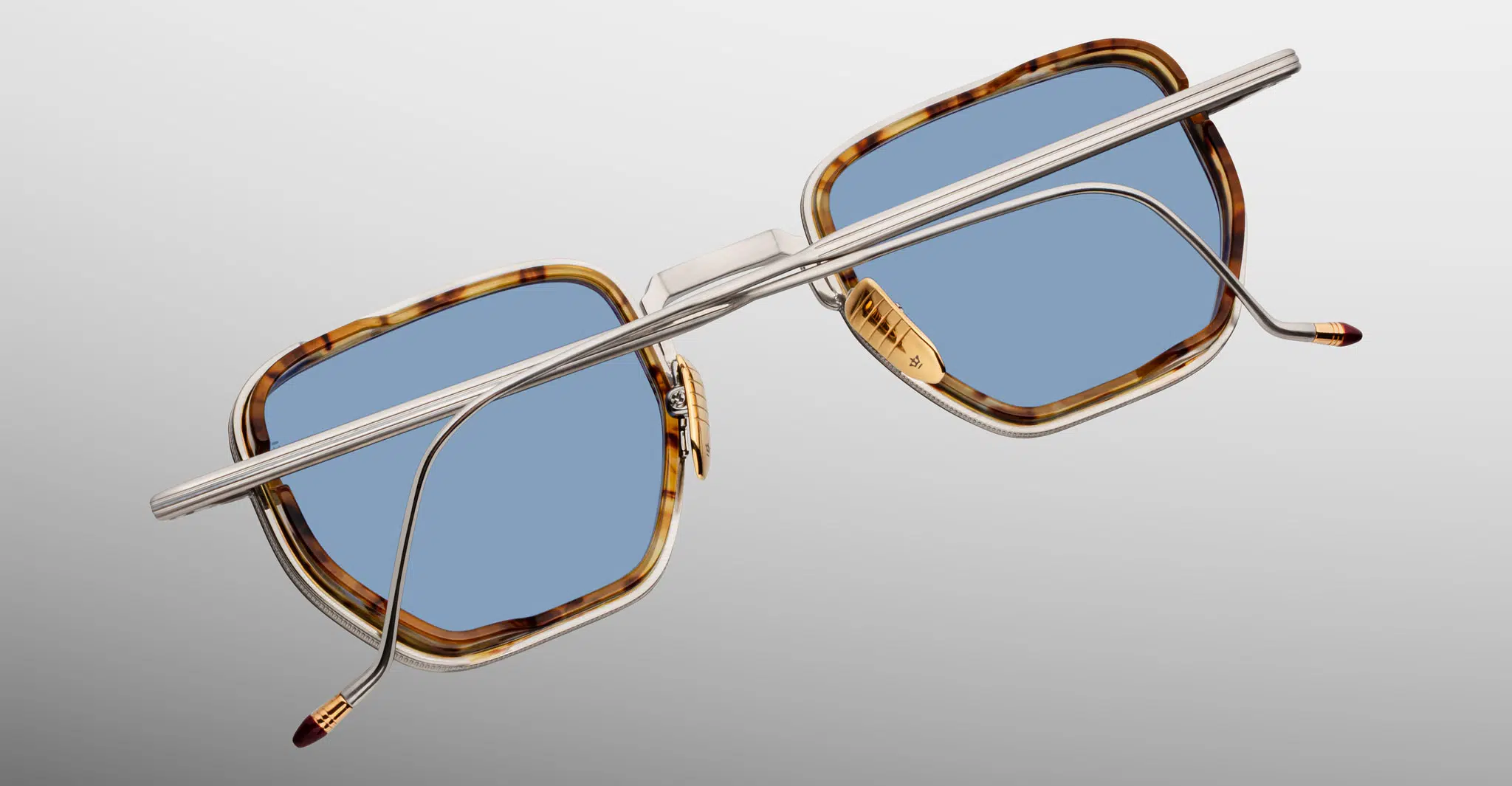 The Jacques Marie Mage Atkins sunglasses feature square light blue lenses, gold and brown tortoiseshell frames, and thin silver metal arms, displayed from the back against a plain gray background.