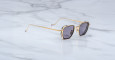 The Jacques Marie Mage Atkins are stylish gold-framed square sunglasses with dark lenses, displayed on a white marble surface.