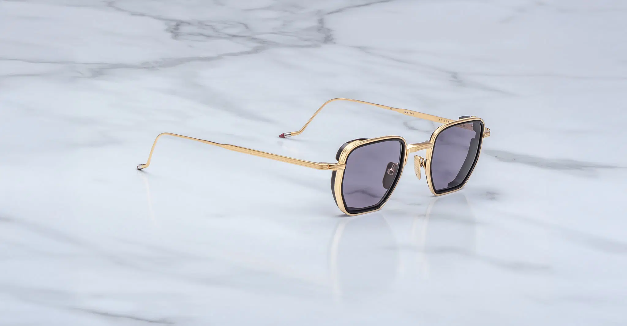 The Jacques Marie Mage Atkins are stylish gold-framed square sunglasses with dark lenses, displayed on a white marble surface.