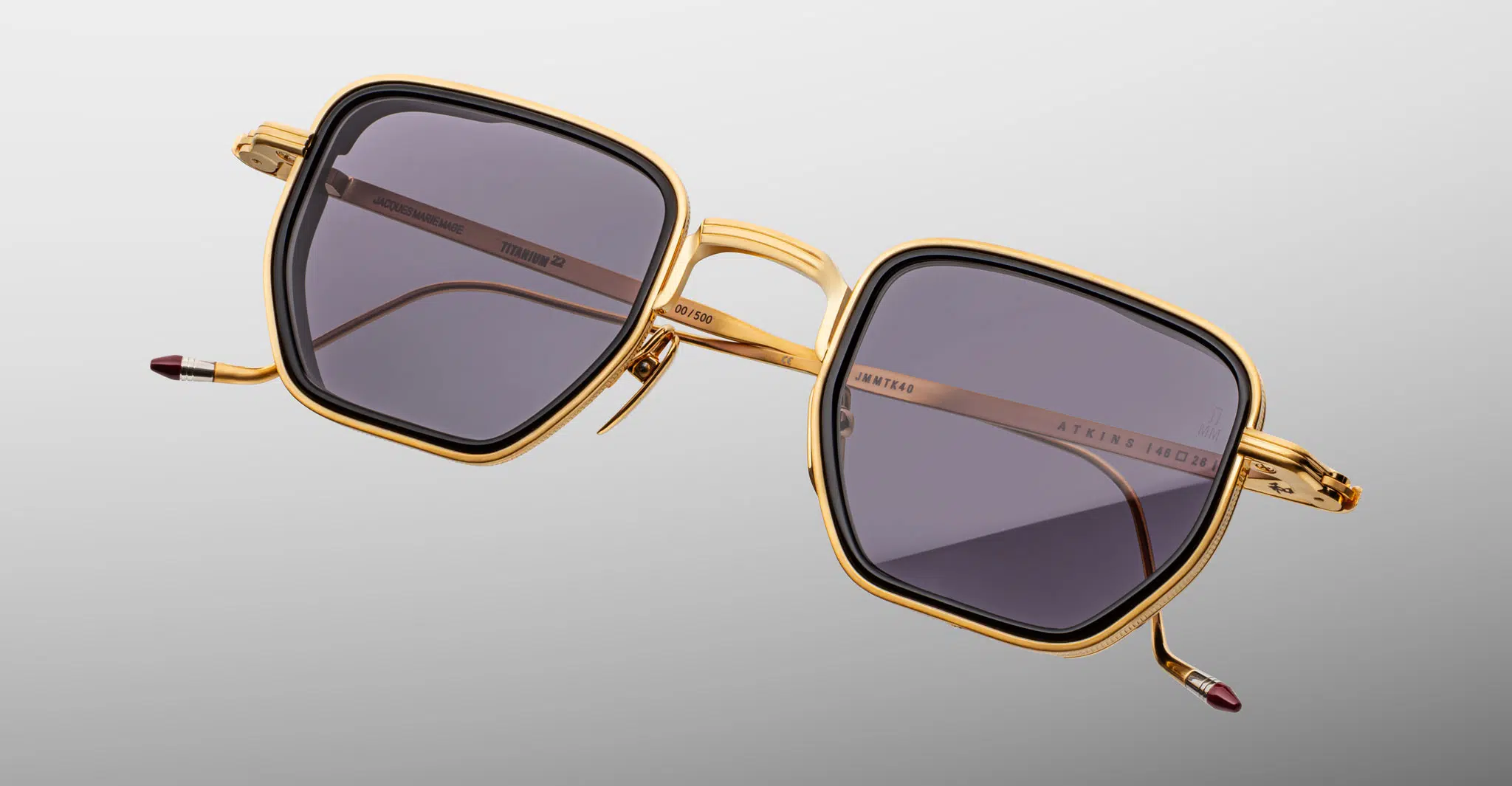 The Jacques Marie Mage Atkins sunglasses, featuring square gold metal frames with dark lenses, are displayed against a plain light gray background. The temples include gold detailing and striking red-tipped ends.