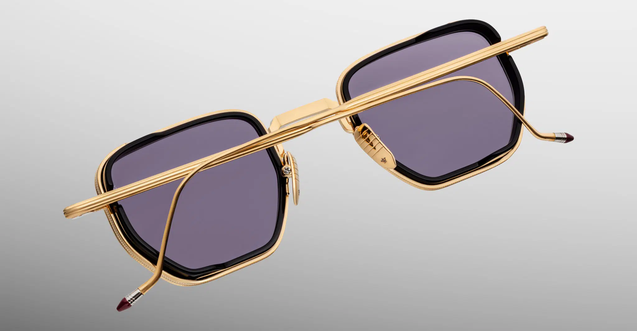 The Jacques Marie Mage Atkins sunglasses feature gold square frames with dark lenses, shown from the back and above to highlight their metallic finish and detailed temples, set against a light gray background.