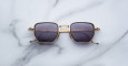 A pair of Jacques Marie Mage Atkins gold-framed square sunglasses with dark tinted lenses is displayed on a white marble surface.