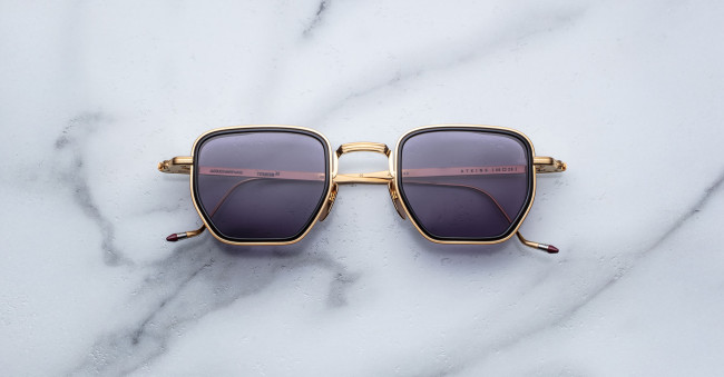 A pair of Jacques Marie Mage Atkins gold-framed square sunglasses with dark tinted lenses is displayed on a white marble surface.