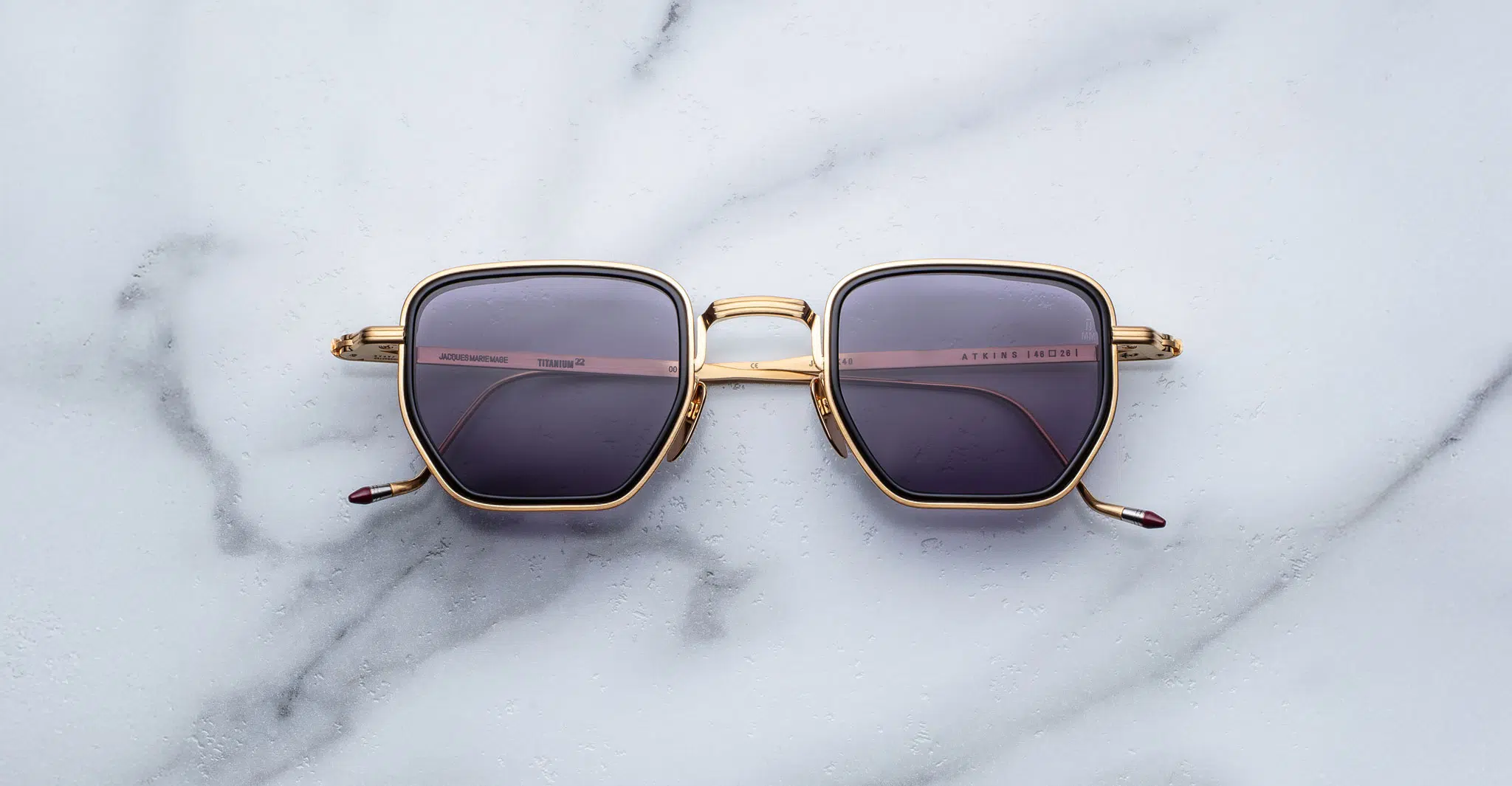 A pair of Jacques Marie Mage Atkins gold-framed square sunglasses with dark tinted lenses is displayed on a white marble surface.