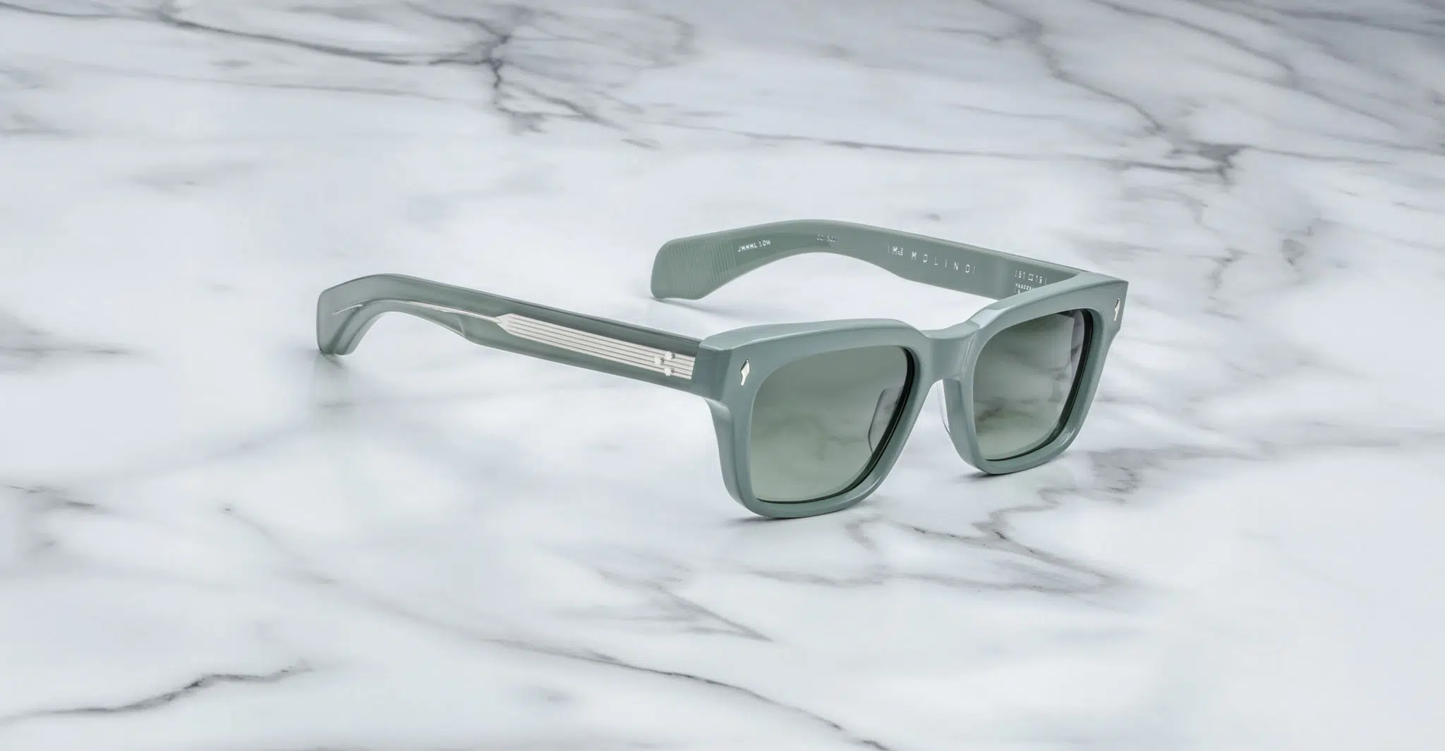 The Jacques Marie Mage Molino sunglasses, featuring mint green square frames, dark lenses, and silver temple details, rest on a white marble surface.