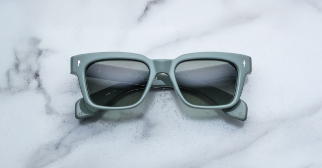 A pair of Jacques Marie Mage Molino sunglasses, featuring a square shape, matte green frames, and dark lenses, rests on a white marble surface.