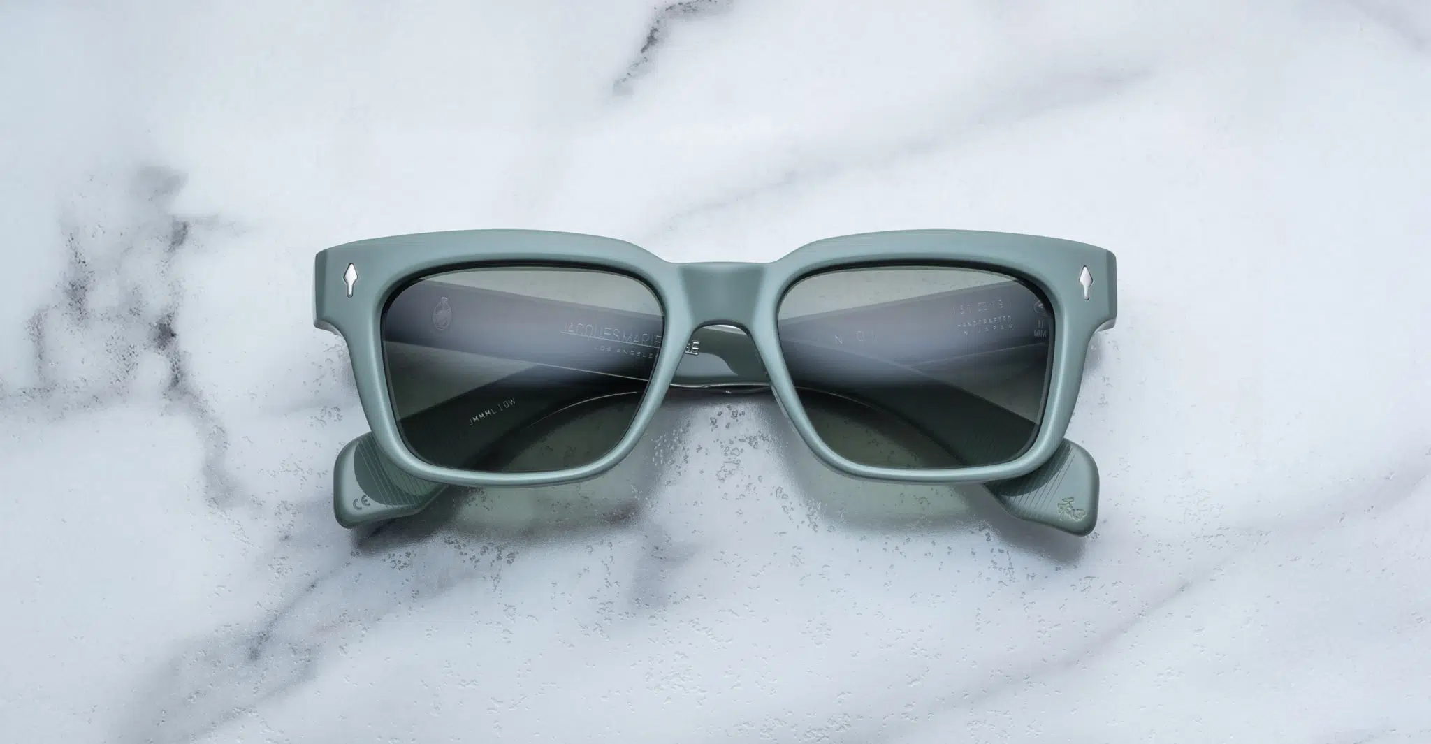 A pair of Jacques Marie Mage Molino sunglasses, featuring a square shape, matte green frames, and dark lenses, rests on a white marble surface.