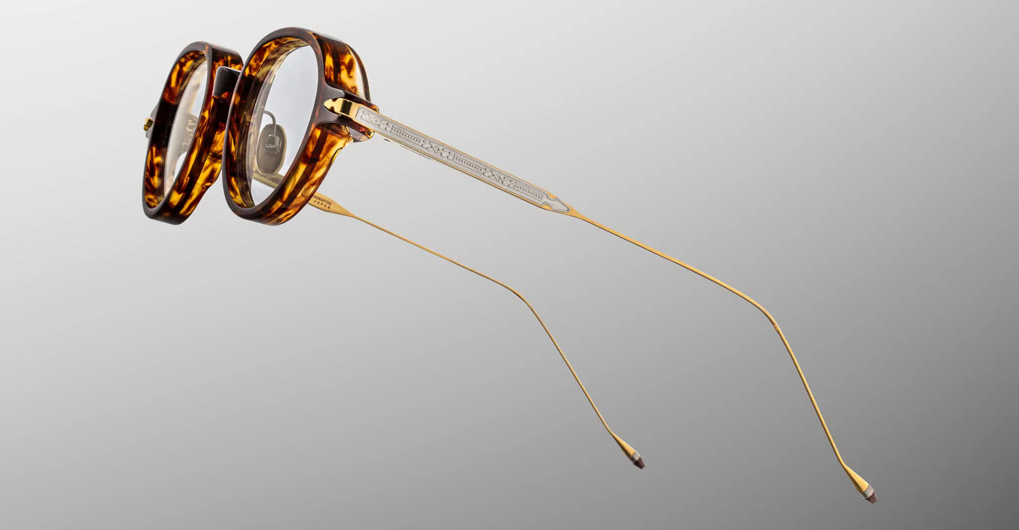 Jacques-Marie-Mage-Clark-Argyle-RX-JMMCRVRX-5C-Detail2-Dan-Deutsch The Jacques Marie Mage Clark eyeglasses feature thick, round tortoiseshell frames with thin gold metal temples, displayed on a light gray gradient background.