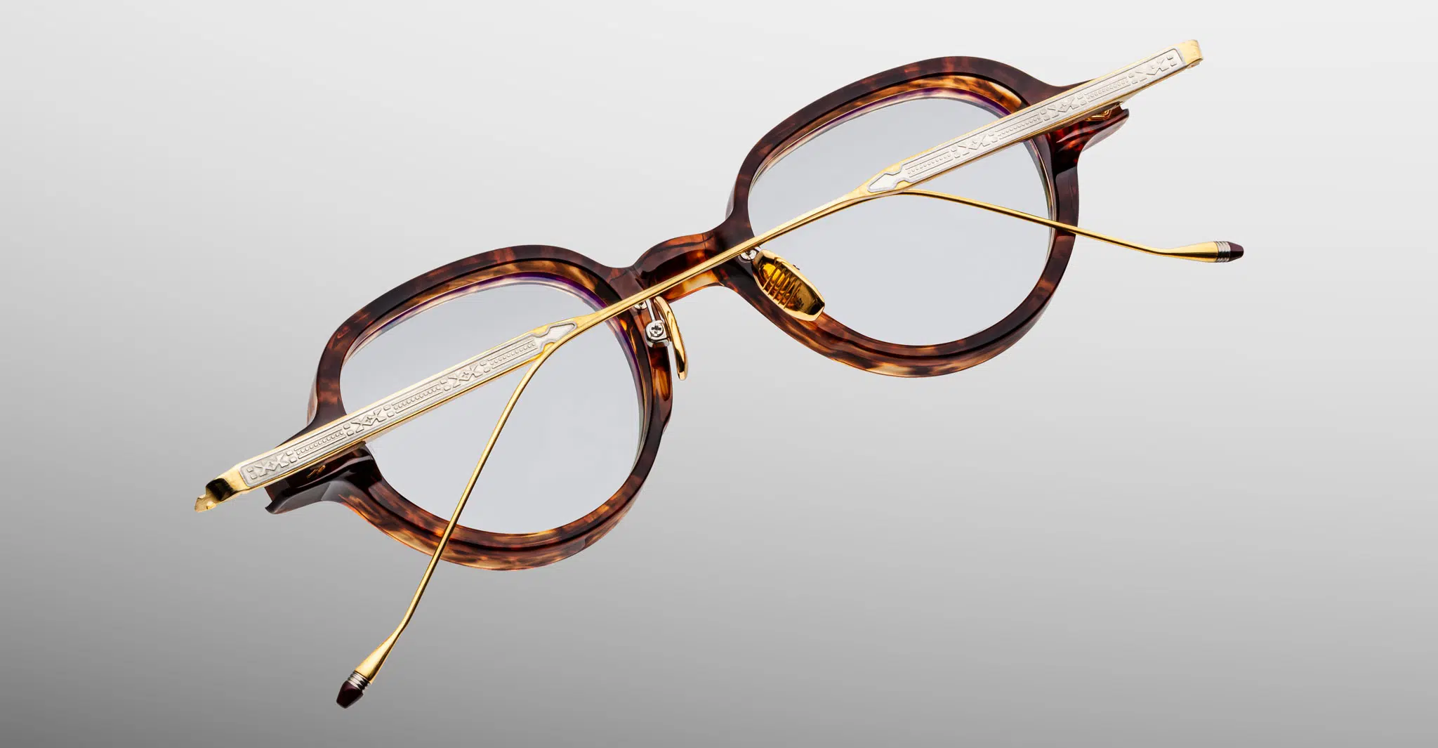 Jacques-Marie-Mage-Clark-Argyle-RX-JMMCRVRX-5C-Detail3-Dan-Deutsch The Jacques Marie Mage Clark eyeglasses feature a tortoiseshell oval frame with gold metal temples and engraved details, displayed from an overhead angle on a light gray background.