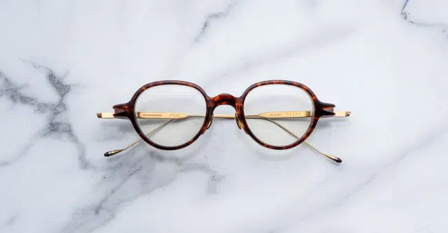 The Jacques Marie Mage Clark, round tortoiseshell eyeglasses with gold temples, are displayed from above on a white marble surface.