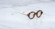 Jacques Marie Mage Clark round tortoiseshell eyeglasses with thin gold metal arms rest on a white marble surface, angled slightly to the right.