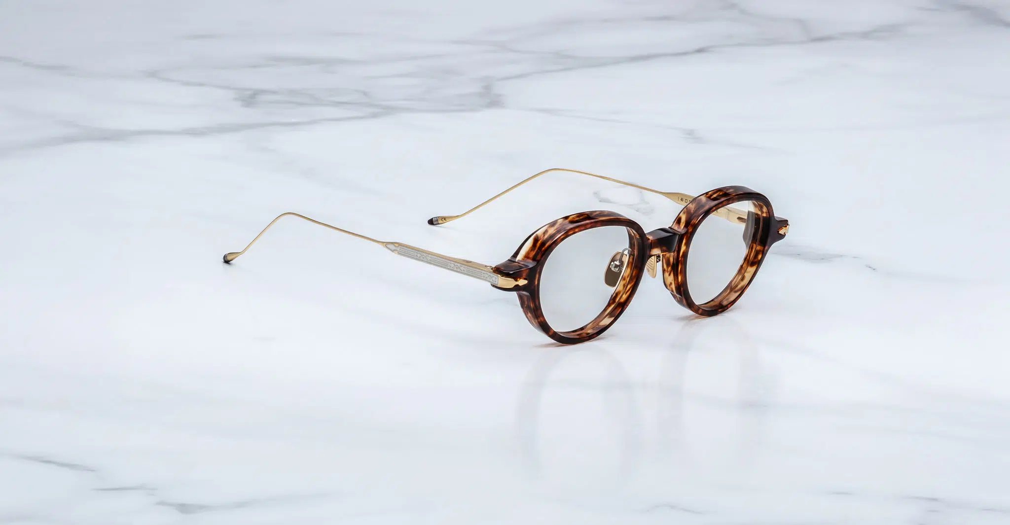 Jacques-Marie-Mage-Clark-Argyle-RX-JMMCRVRX-5C-Profile-Dan-Deutsch Jacques Marie Mage Clark round tortoiseshell eyeglasses with thin gold metal arms rest on a white marble surface, angled slightly to the right.