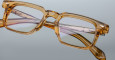 Close-up of Jacques Marie Mage Moscova eyeglasses, featuring thick rectangular translucent amber frames with visible hinges, engraved temples, and metal details, set against a plain gray background.