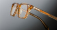Close-up of Jacques Marie Mage Moscova eyeglasses featuring thick, transparent amber-colored frames and metal hinges, photographed against a gradient gray background.