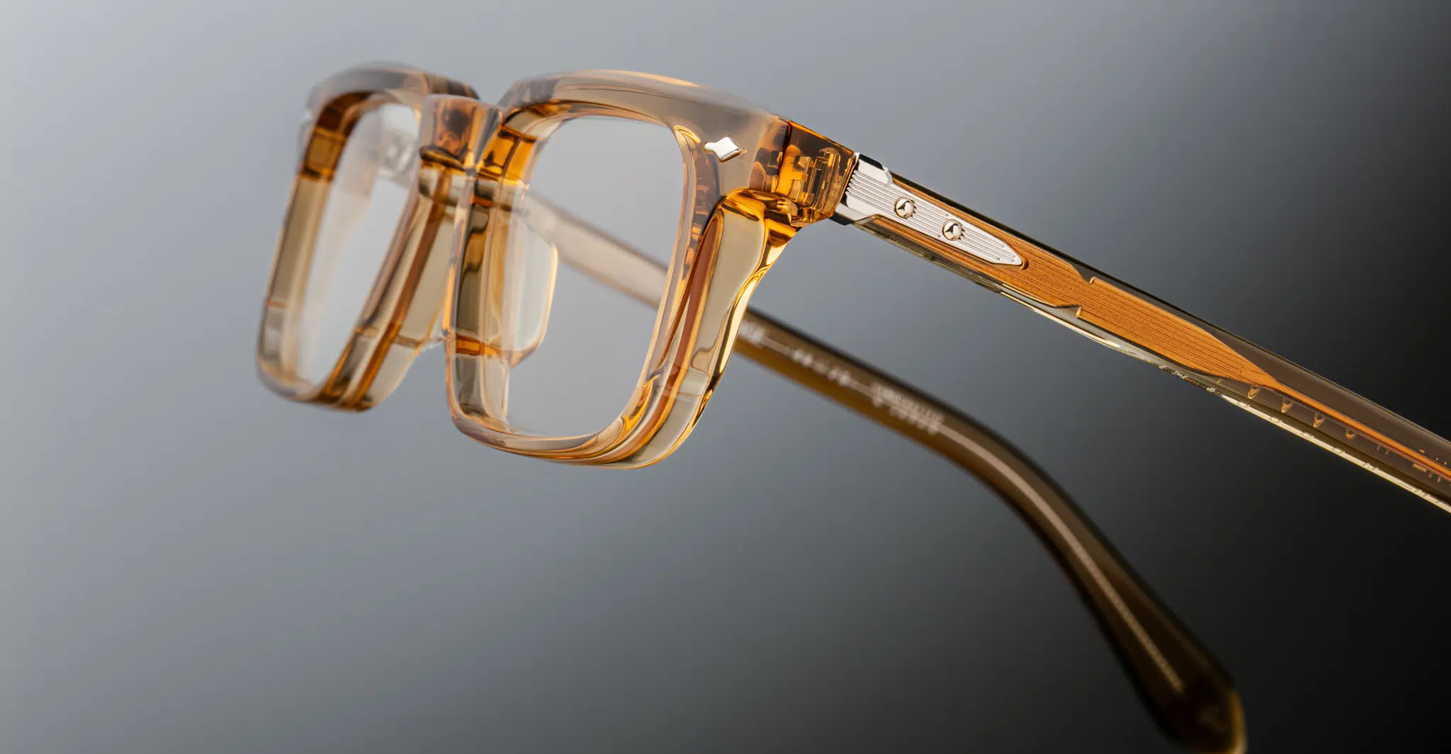 Close-up of Jacques Marie Mage Moscova eyeglasses featuring thick, transparent amber-colored frames and metal hinges, photographed against a gradient gray background.