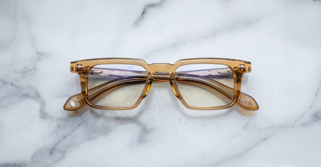 A pair of Jacques Marie Mage Moscova translucent brown rectangular eyeglasses rests on a white marble surface with gray veining, arms folded inward and lenses facing up.