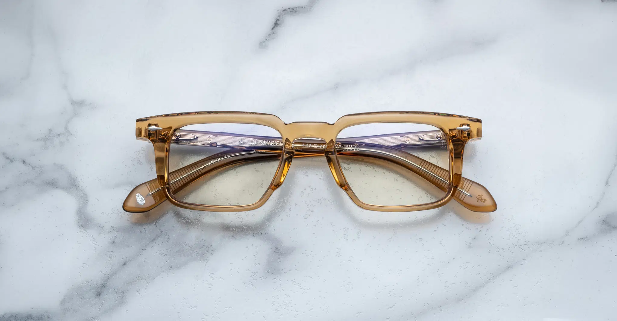 A pair of Jacques Marie Mage Moscova translucent brown rectangular eyeglasses rests on a white marble surface with gray veining, arms folded inward and lenses facing up.