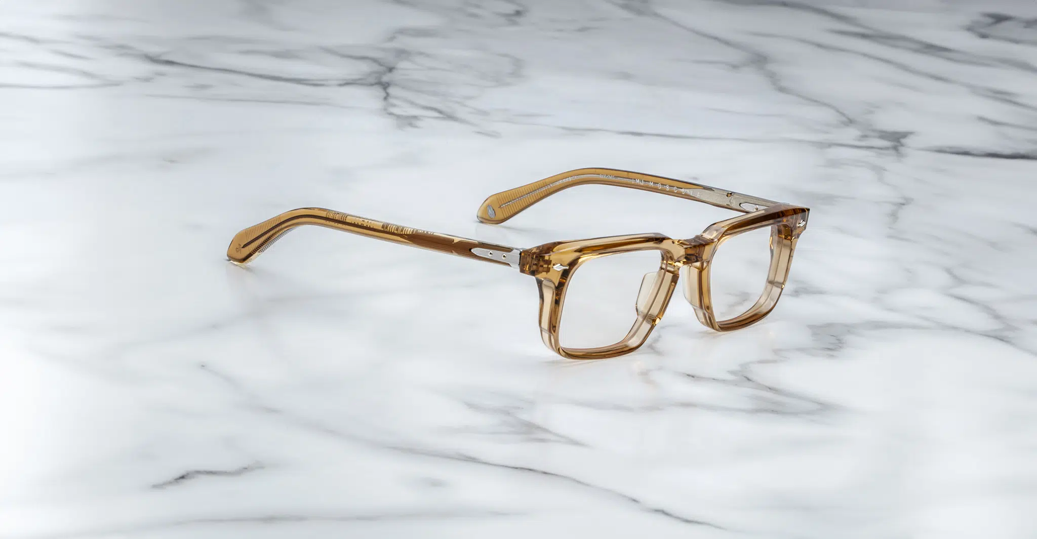The Jacques Marie Mage Moscova brown transparent square-frame eyeglasses are displayed on a white marble surface with gray veining.
