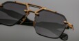 A close-up of Jacques Marie Mage Silverton black sunglasses with dark tinted lenses and gold metallic accents on the frame and bridge, set against a plain gray background.