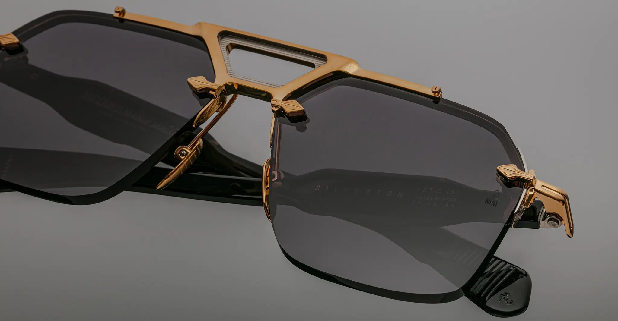 A close-up of Jacques Marie Mage Silverton black sunglasses with dark tinted lenses and gold metallic accents on the frame and bridge, set against a plain gray background.