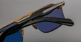 Close-up view of Jacques Marie Mage Silverton sunglasses featuring gold frames, blue-tinted lenses, and black arms, set against a plain gray background.