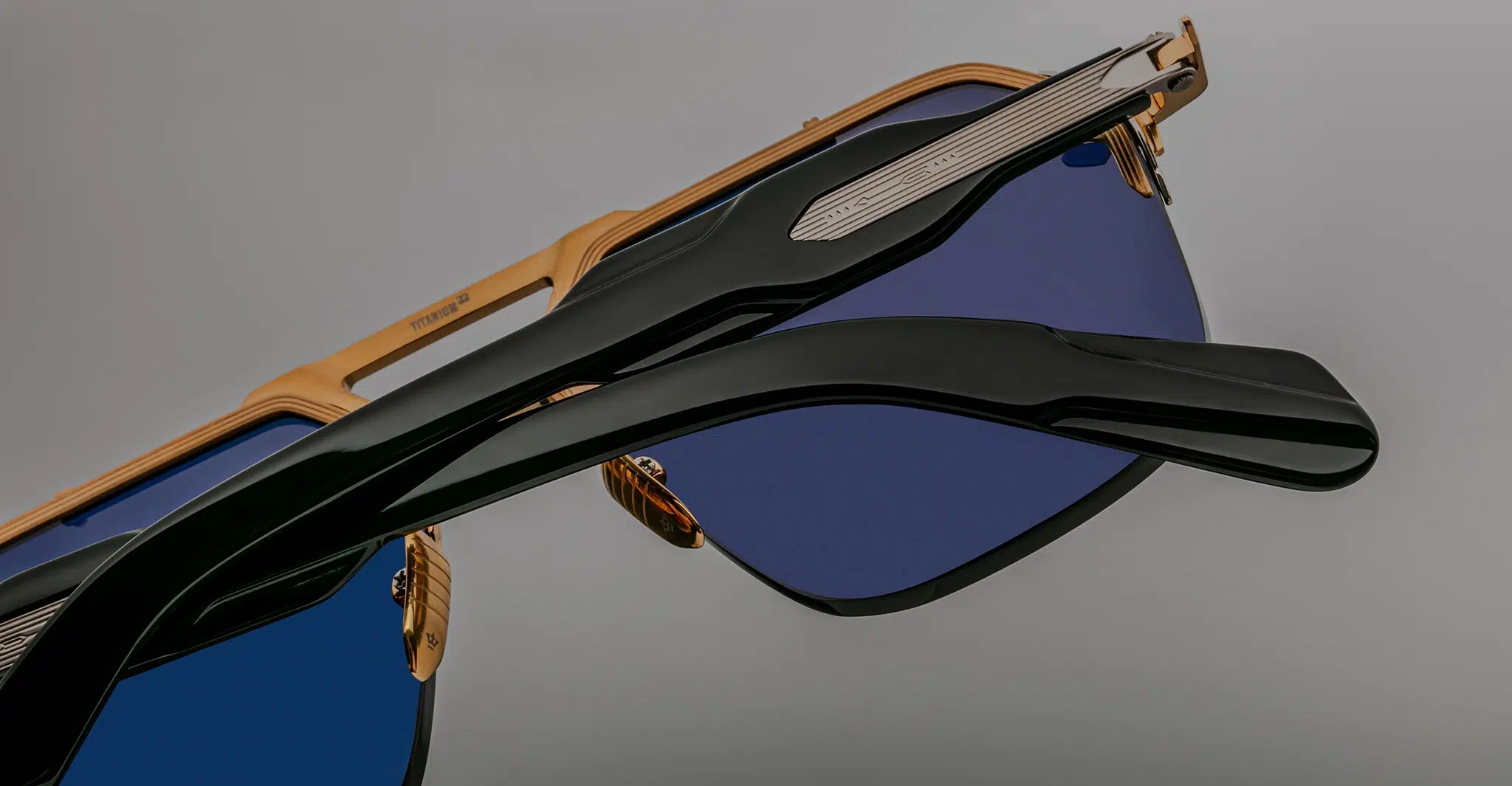 Close-up view of Jacques Marie Mage Silverton sunglasses featuring gold frames, blue-tinted lenses, and black arms, set against a plain gray background.