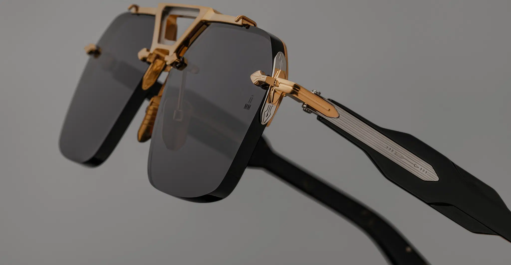 Close-up of Jacques Marie Mage Silverton sunglasses with dark rectangular lenses, gold metal frame, and black arms with metallic detailing, set against a plain gray background.