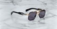 The Jacques Marie Mage Silverton sunglasses feature dark rectangular lenses, gold-tone metal frames, and thick black arms, displayed on a white marble surface with grey veining.