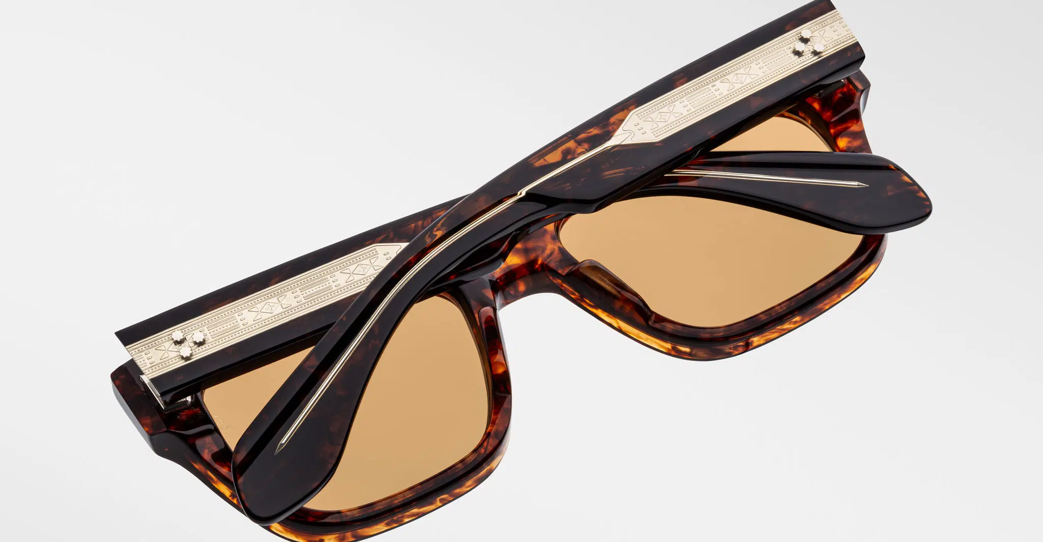 A pair of Jacques Marie Mage Cash tortoiseshell sunglasses with brown lenses, highlighted by gold-toned decorative metal accents on the arms and top edge, is shown folded against a plain light background.