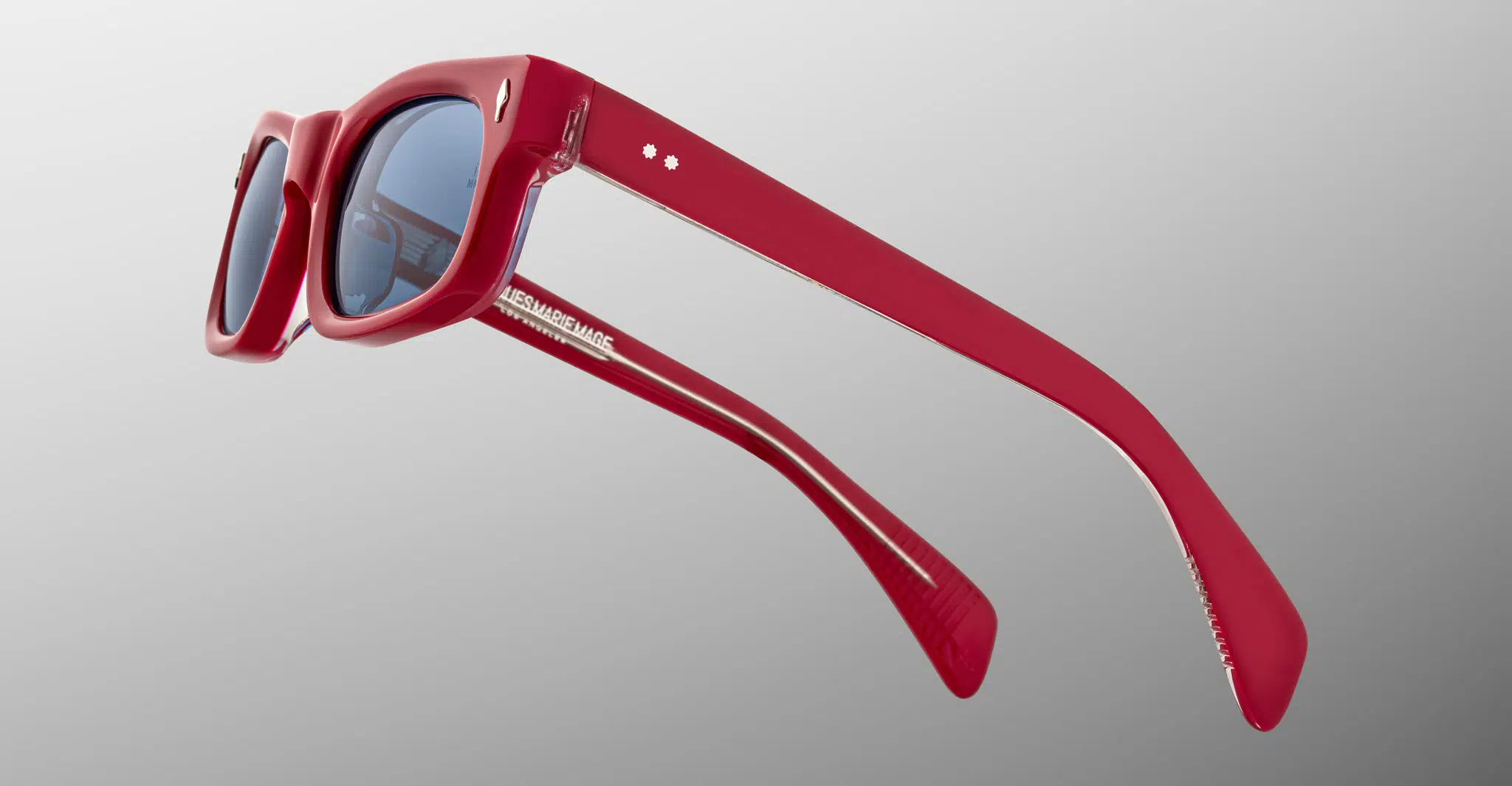 The Jacques Marie Mage Initials red acetate sunglasses feature thick frames, dark lenses, and white printed branding on the inside arm. Shown from a side angle against a light gray gradient background.