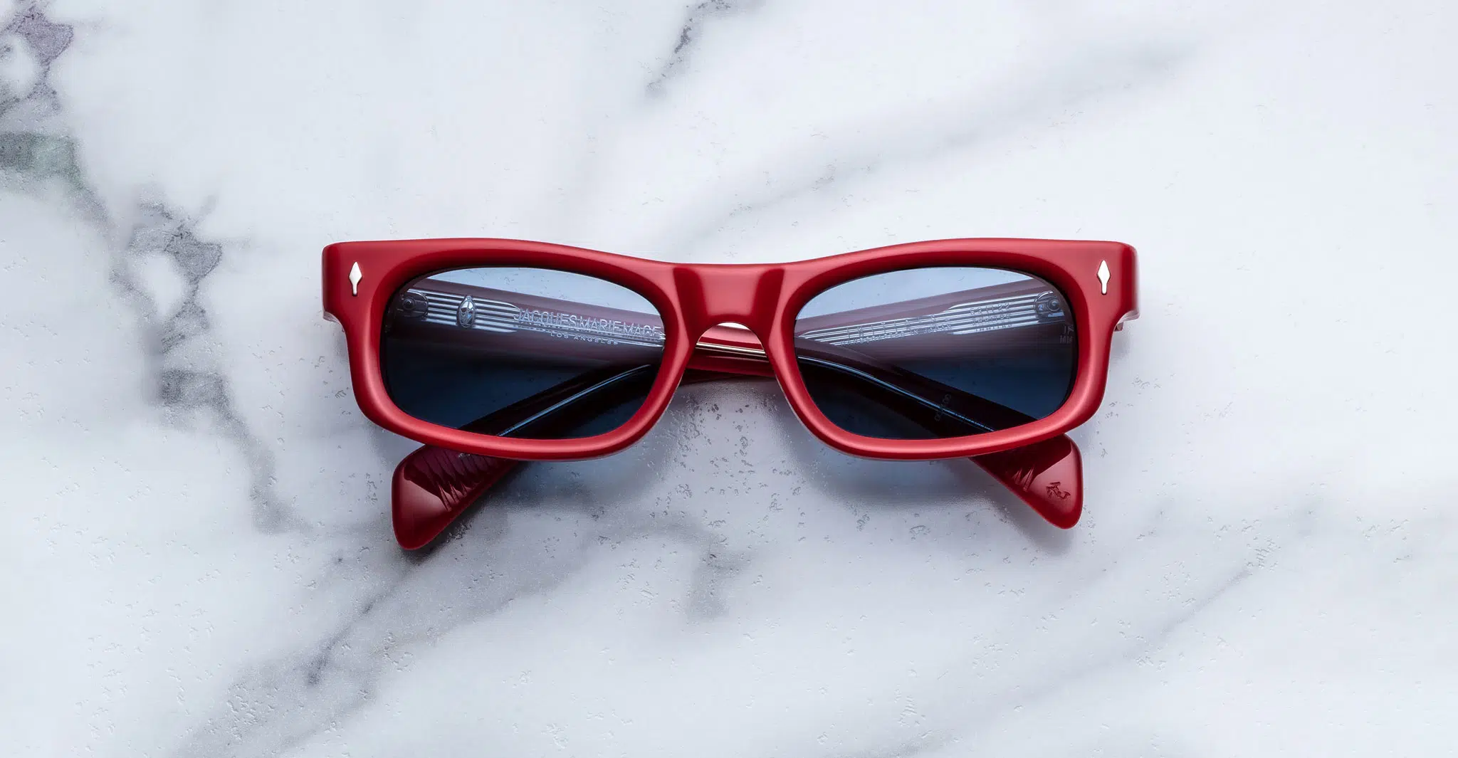 Jacques Marie Mage Initials sunglasses featuring red frames and dark lenses, folded neatly on a white marble surface with subtle gray veins.
