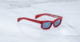 A pair of Jacques Marie Mage Initials sunglasses with red frames and dark lenses sits on a white marble surface.