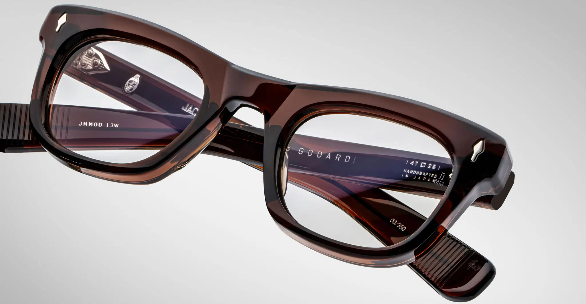 A close-up of rectangular, thick-framed Jacques Marie Mage Godard brown eyeglasses with translucent lenses on a plain gray background. The brand name and model details appear on the inside of the frame arms.