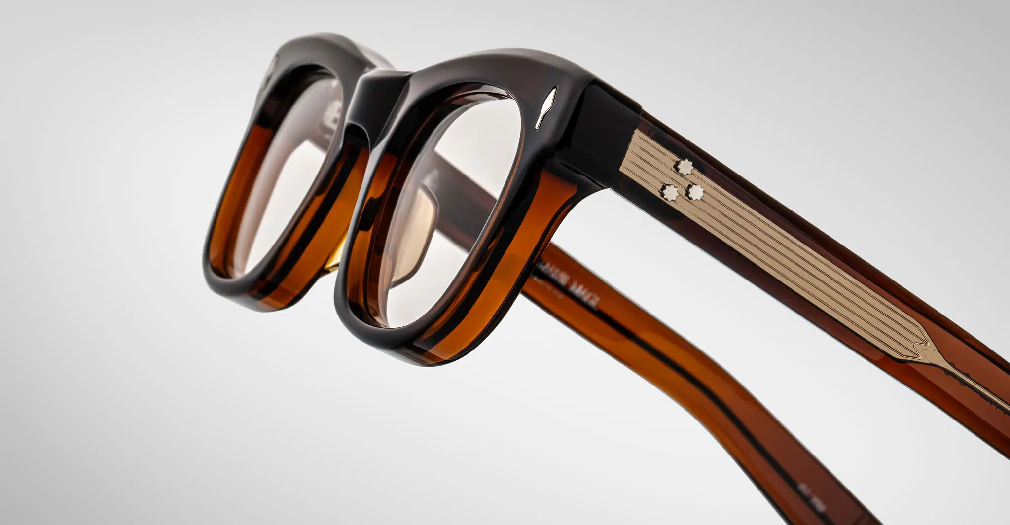 Close-up of Jacques Marie Mage Godard eyeglasses featuring brown and black thick frames, transparent lenses, and metal temple accents, displayed against a plain light background.