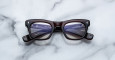 A pair of Jacques Marie Mage Godard dark-rimmed rectangular eyeglasses with clear lenses rests on a white marble surface, showing the brand name and specifications on the inside of the temple arms.