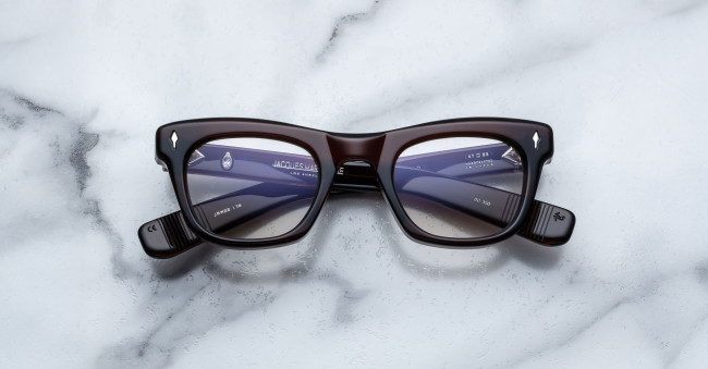 A pair of Jacques Marie Mage Godard dark-rimmed rectangular eyeglasses with clear lenses rests on a white marble surface, showing the brand name and specifications on the inside of the temple arms.