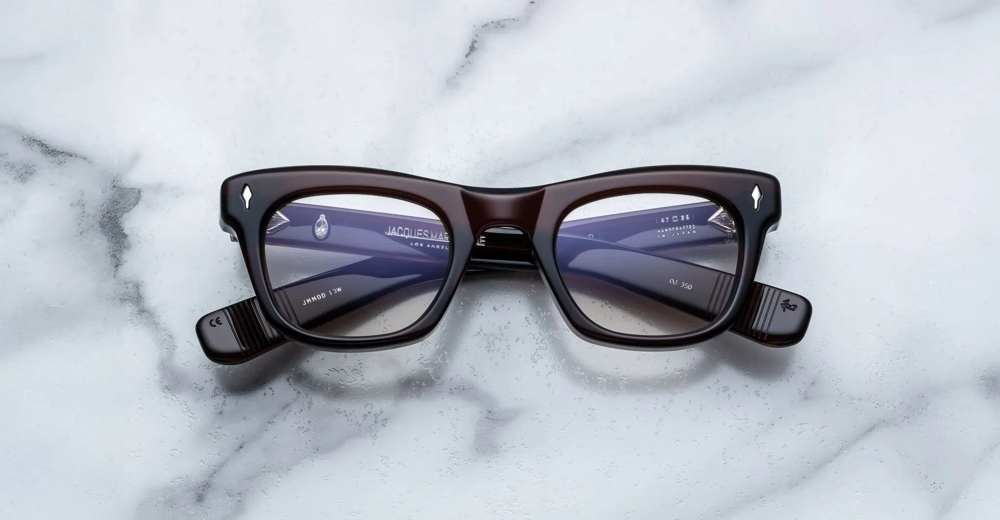 A pair of Jacques Marie Mage Godard dark-rimmed rectangular eyeglasses with clear lenses rests on a white marble surface, showing the brand name and specifications on the inside of the temple arms.