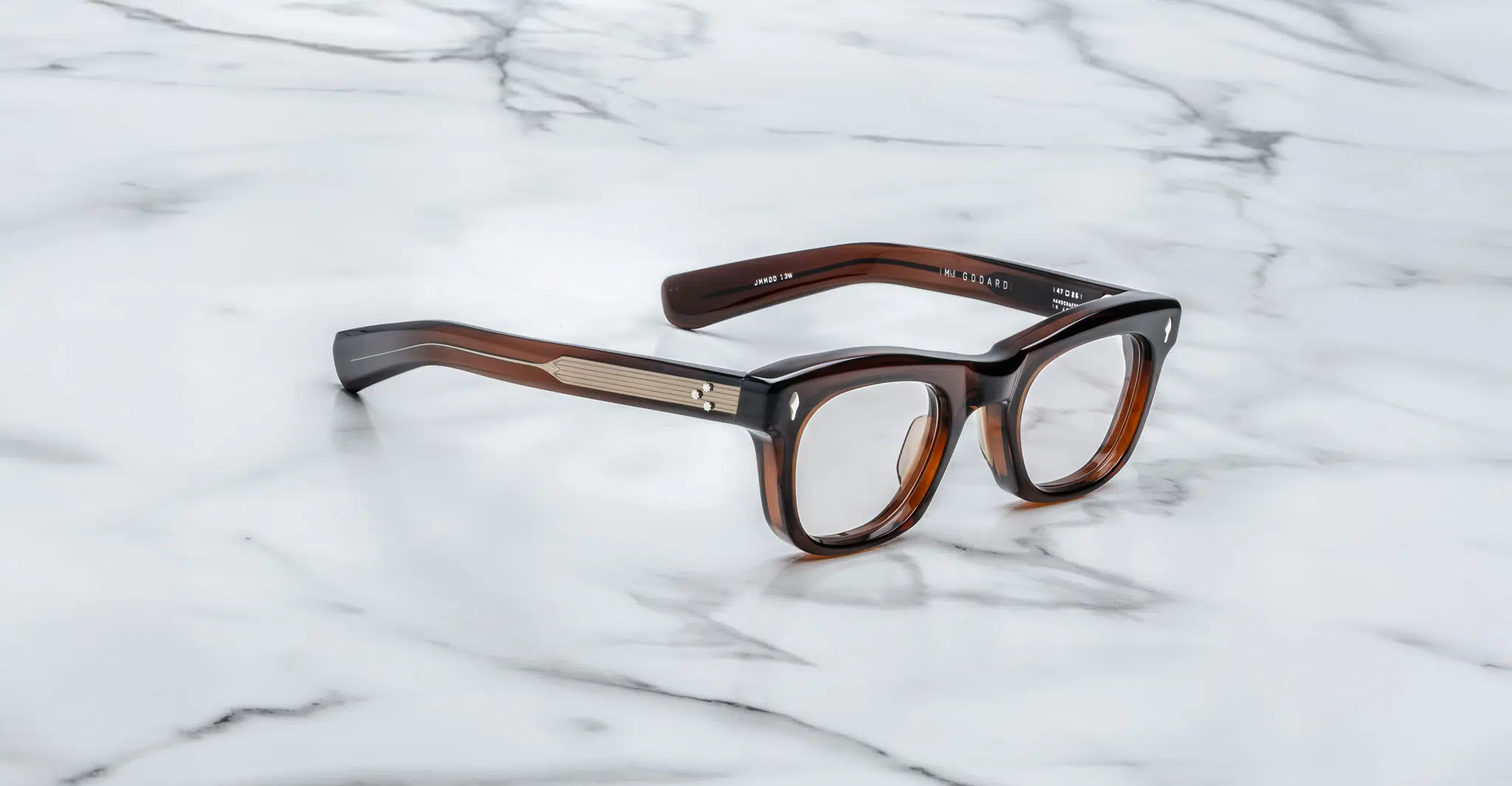 The Jacques Marie Mage Godard eyeglasses feature thick brown tortoiseshell rectangular frames with gold detailing on the temples, displayed on a white marble surface.