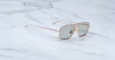 The Jacques Marie Mage Earl sunglasses, featuring gold frames and green-tinted rectangular lenses, rest on a white marble surface with gray veining.