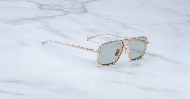 The Jacques Marie Mage Earl sunglasses, featuring gold frames and green-tinted rectangular lenses, rest on a white marble surface with gray veining.