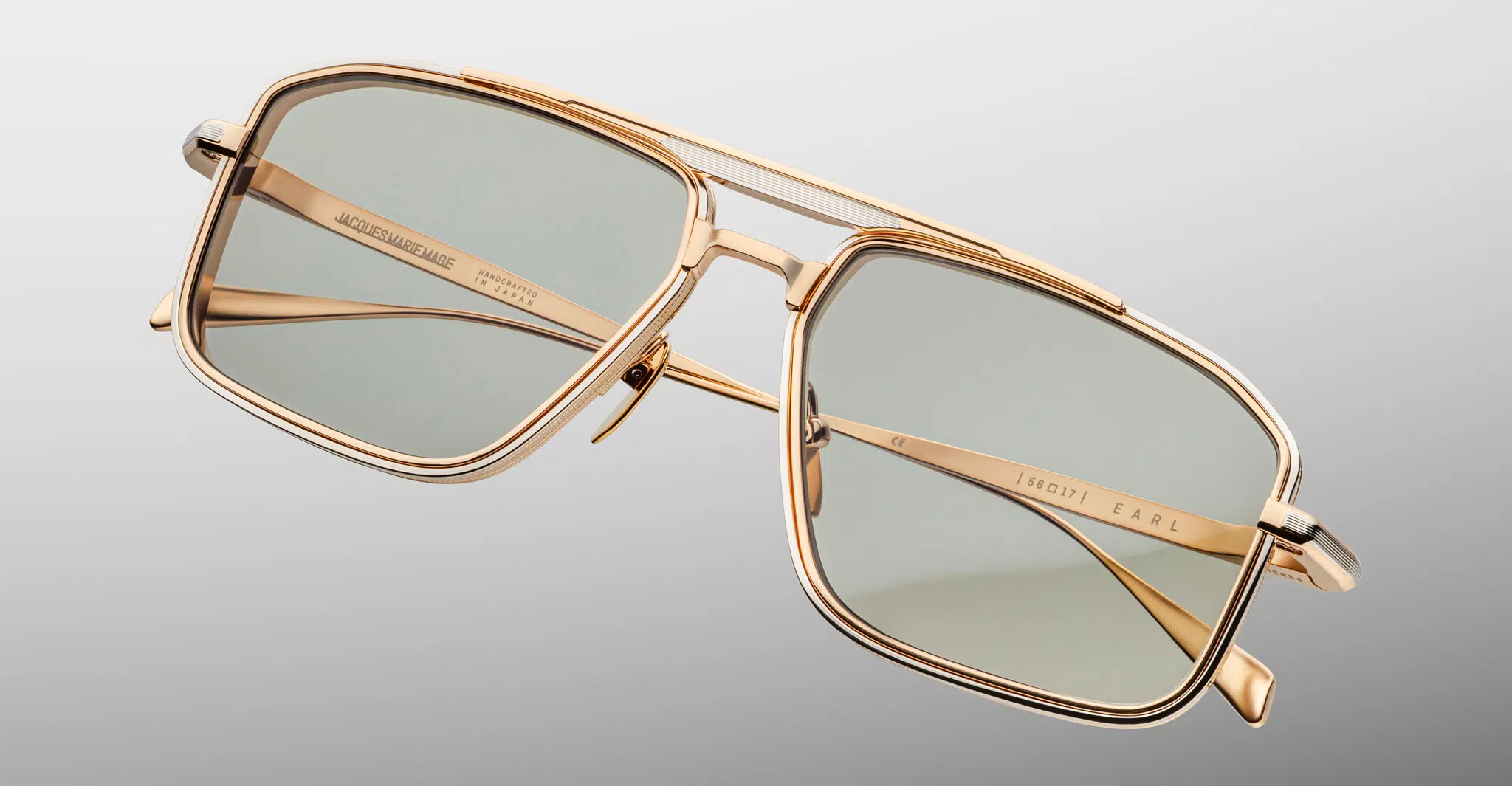 The Jacques Marie Mage Earl sunglasses feature a gold metal frame with rectangular green-tinted lenses, shown at an angle on a light gray background. The double bridge design and engraved branding on the arms complete the look.